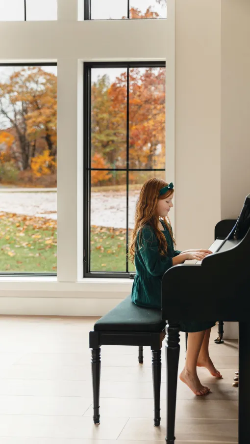 Young girl in green dress playing piano barefoot beside large windows with autumn trees outside.
