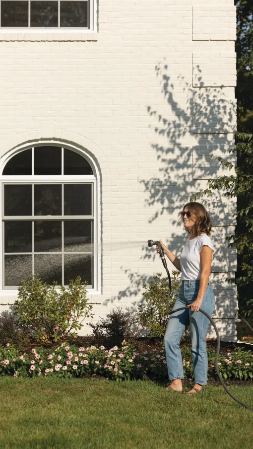 Woman in sunglasses watering flower garden with hose in front of white brick house on a sunny day
