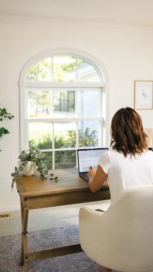 Woman working on a laptop at a wooden desk in a bright home office with a large window and indoor plants.