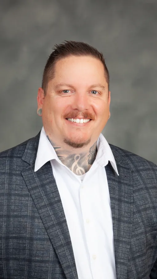 Smiling man in a checkered suit jacket and white shirt with a neck tattoo and short hair against a gray backdrop