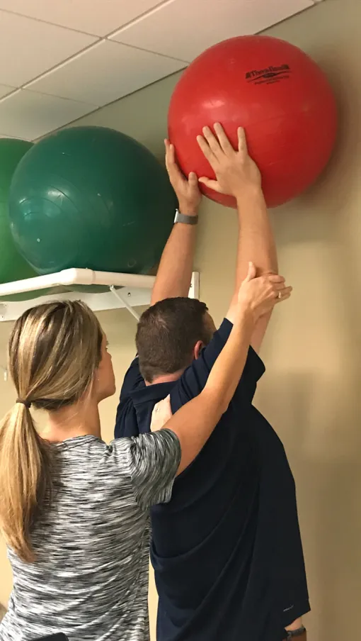 Physical therapist assisting a man reaching for a red exercise ball on a wall shelf during rehabilitation.