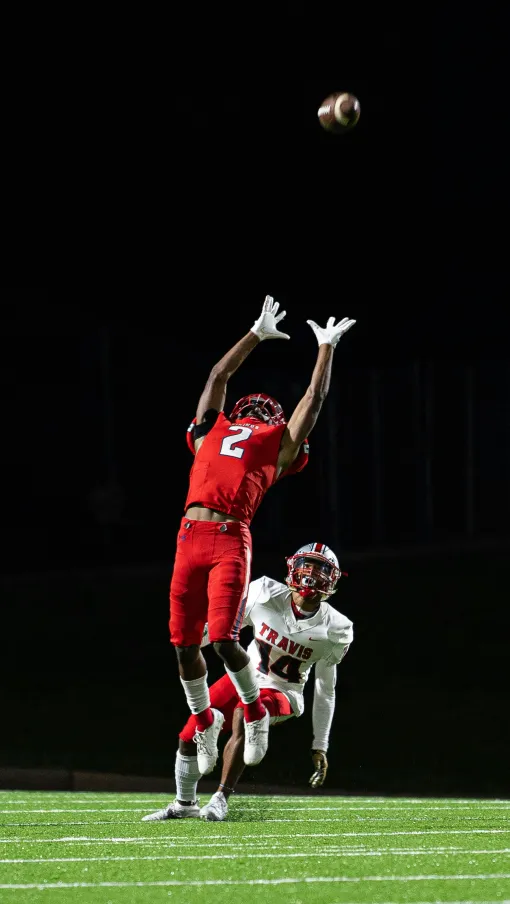 Football player in red jersey jumps to catch a high pass under stadium lights during night game.