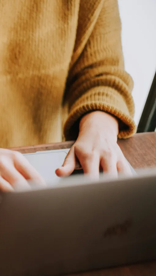 Person wearing a mustard sweater typing on a laptop at a wooden table with bracelets on wrist