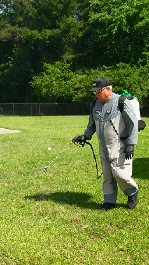Person in protective gear spraying pesticide on green lawn near patio with chairs on sunny day.