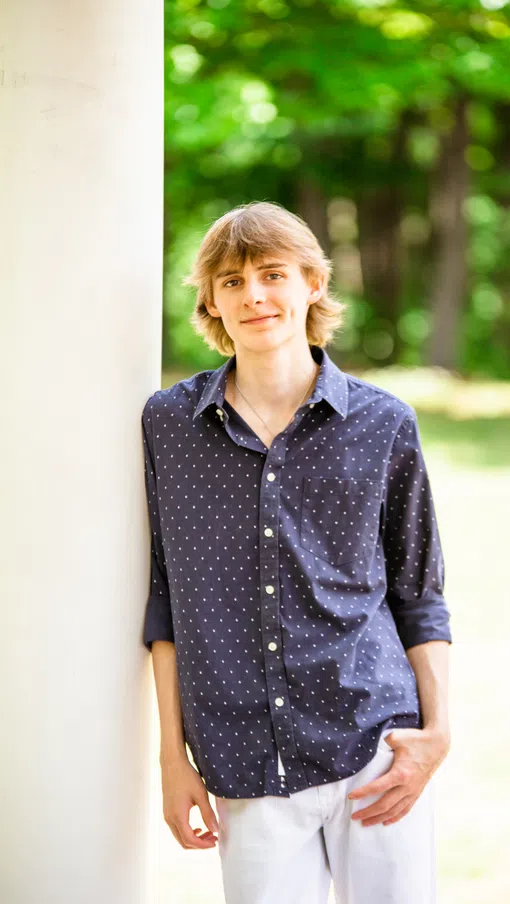 Young man in navy polka dot shirt and white pants leans against a white pillar in a green outdoor setting