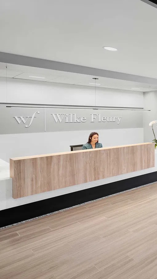 Modern office reception area with wooden desk, seating, and employees working in a bright space.