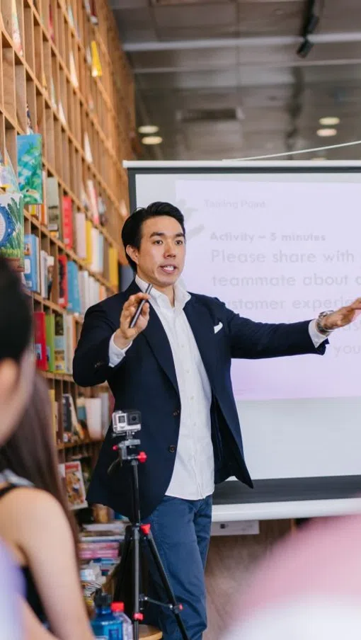 Young man giving a presentation in a library with audience and a projection screen in the background