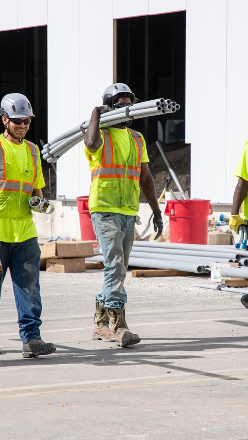Four construction workers in safety gear carry PVC pipes while walking on a construction site outdoors.