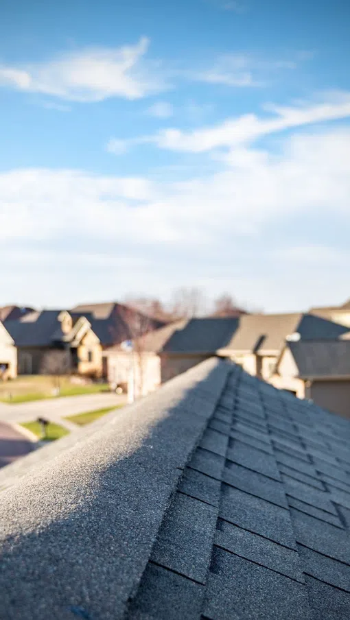 Close-up of a gray shingled roof with a suburban neighborhood and blue sky in the background