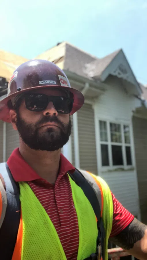 Construction worker in safety vest and helmet stands in front of a house with roofers working in the background.