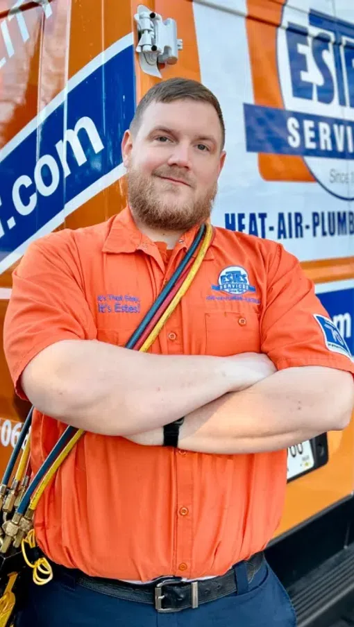 HVAC technician in orange uniform standing confidently with tools across chest in front of Estes Services van.