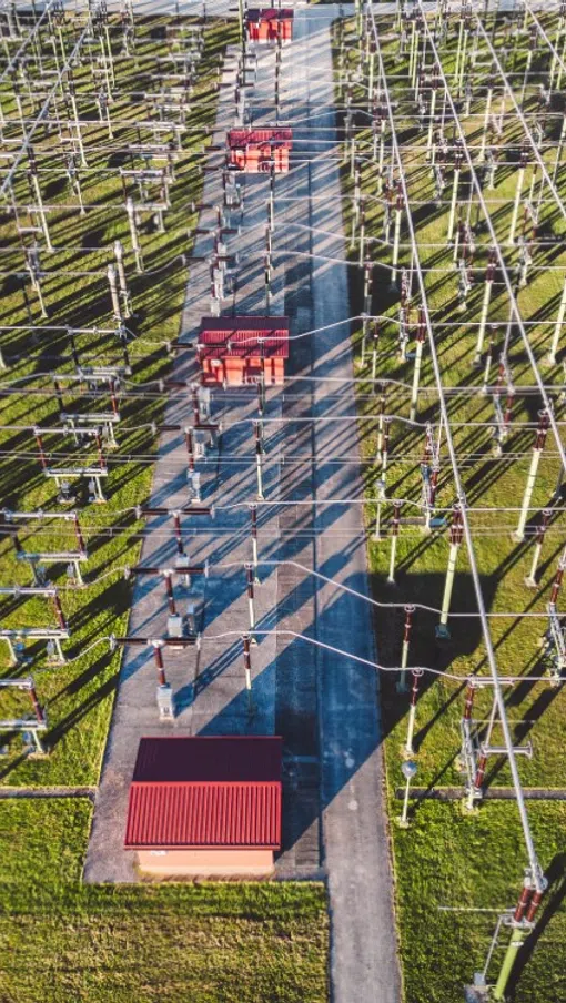 Aerial view of an electrical substation with metal frameworks and red equipment sheds casting long shadows on green grass.