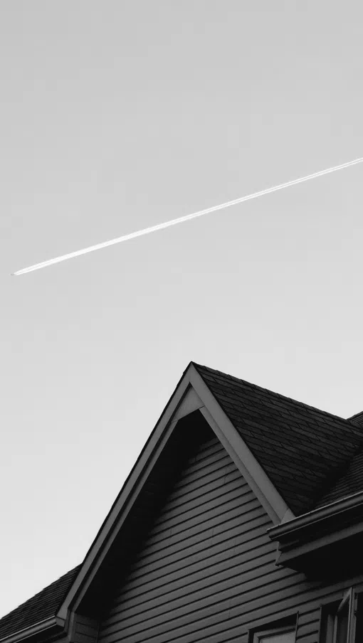 Black and white photo of a house roof with a clear sky and a plane's contrail above it.