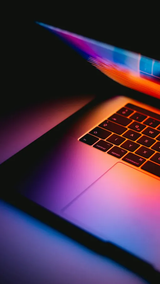 Close-up of a laptop keyboard and partially open screen illuminated by colorful purple and orange lights.