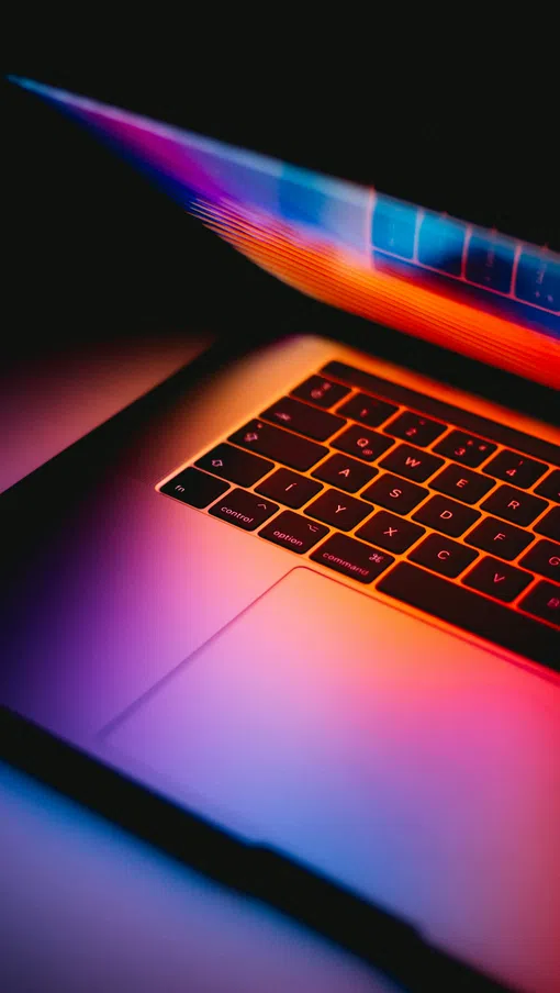 Partially open laptop keyboard illuminated by vibrant purple, orange, and blue lighting in a dark setting