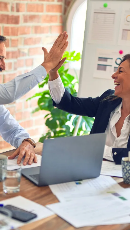 Two business colleagues smiling and giving a high-five in a bright modern office with paperwork and laptop on desk.