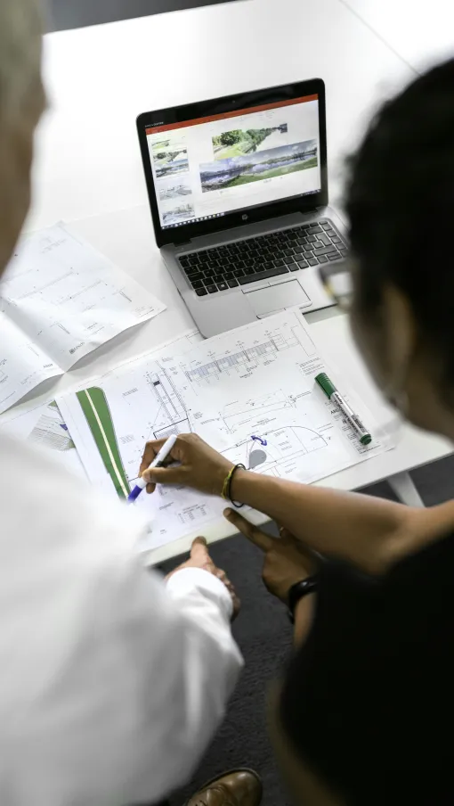 Two people reviewing architectural blueprints and design plans with a laptop showing landscape visuals.