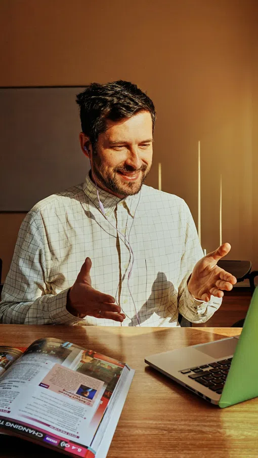 Man with earbuds smiling and gesturing during a video call on a laptop in a sunlit office.