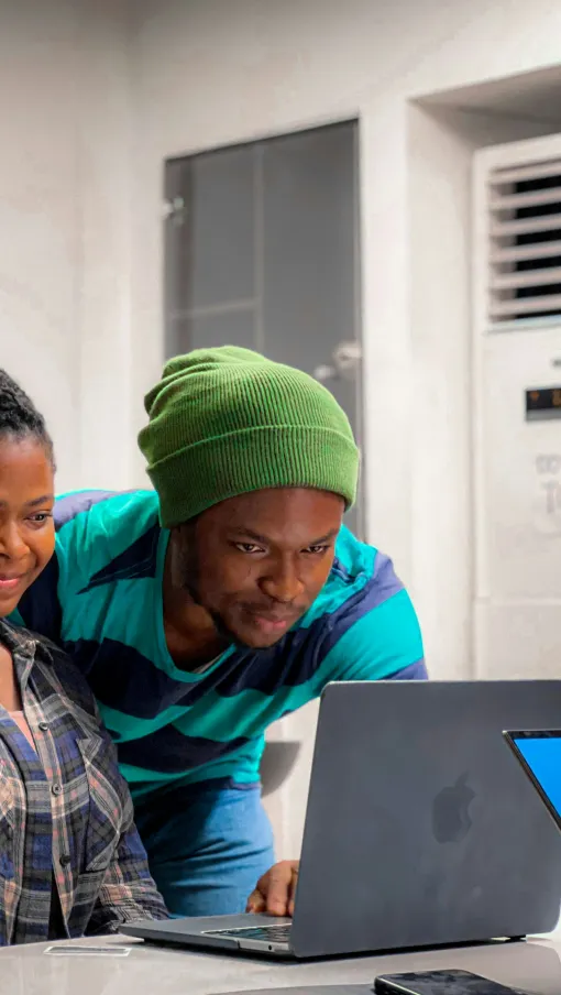 Two young adults smiling and looking at a laptop screen together in a bright room near a table.