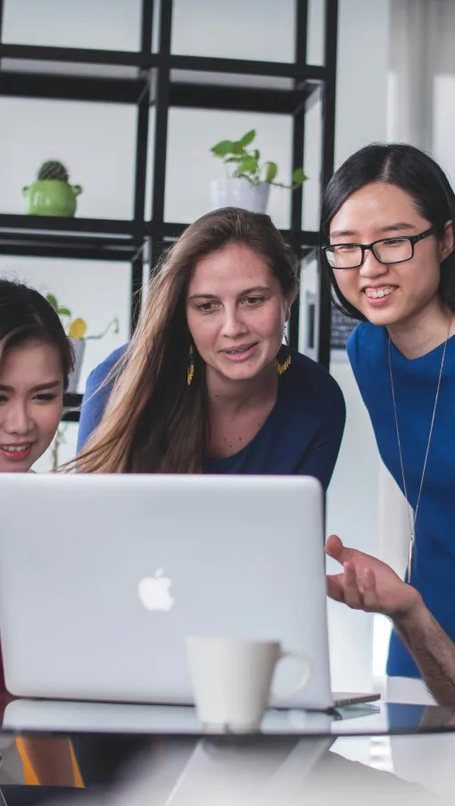 Four diverse colleagues collaborating around a laptop in a modern office setting with plants and a coffee cup.