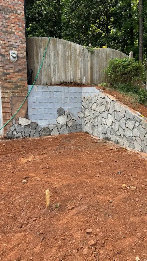 Stone retaining wall under construction in a landscaped outdoor area with wooden fence and tools visible.