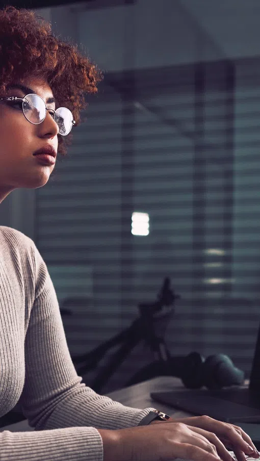 Focused woman with curly hair and glasses typing on keyboard at a desk with laptop in a modern office.