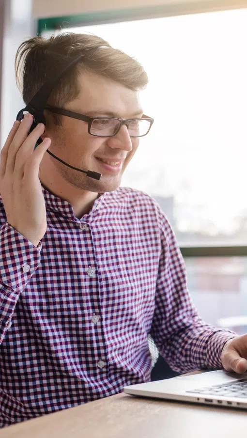 Young man with headset working on laptop in bright office, multitasking with computer and communication device.