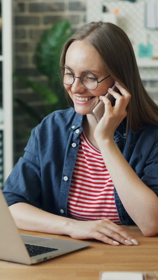 Young woman in glasses happily talking on phone while working on laptop at wooden desk indoors.