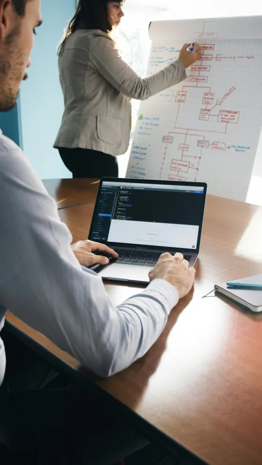 Man coding on laptop while woman writes on flowchart on whiteboard in office meeting.
