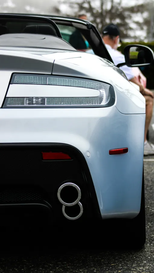 Close-up of a silver sports car's rear with distinctive taillight, dual exhaust, and sleek design in a parking lot.