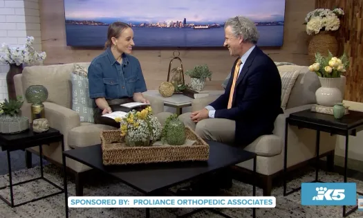 Two hosts sitting and chatting on a cozy TV show set with floral decor and cityscape backdrop.