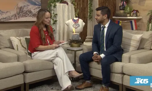 Woman in red shirt interviewing man in suit on beige sofas with Olympic torch and medals in background.
