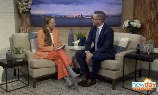 TV hosts seated and conversing on New Day Northwest set with cityscape backdrop and cozy studio decor..