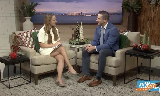Two people sitting in a cozy studio set decorated with holiday ornaments and a city skyline backdrop.