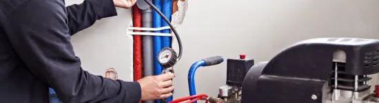 Technician checking water pressure gauges on pipes connected to a blue air compressor in a wall cavity.