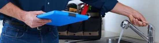 Man in blue shirt checking water flow from kitchen sink faucet with clipboard in hand and tools nearby.