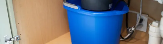 Blue bucket placed under kitchen sink pipes and garbage disposal unit inside wooden cabinet.
