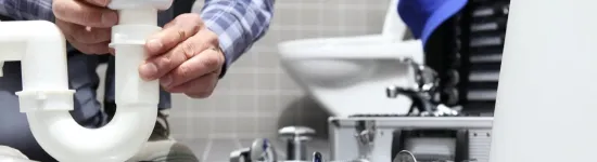 Plumber assembling white PVC pipe under a sink amid plumbing tools and parts in a tiled bathroom.