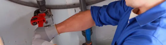 Plumber in blue uniform and hard hat repairing pipe under sink with red pipe wrench and gloves