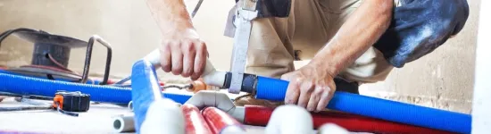 Plumber working with blue and red pipes using a cutting tool on a concrete floor in construction site.