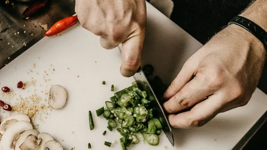 Chef slicing green okra on a white cutting board with mushrooms, salmon, and chili peppers nearby.
