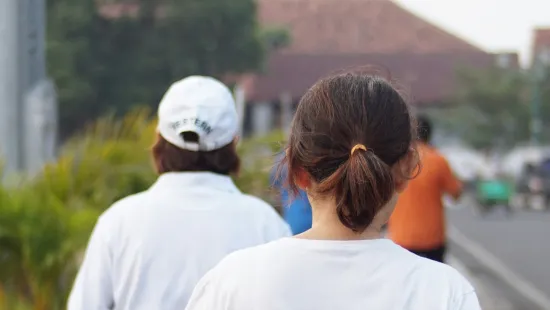 Two people walking outdoors on a sidewalk near greenery and buildings, seen from behind in casual clothes.