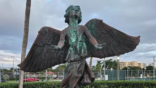 Bronze angel statue with wings spread wide in outdoor park near palm trees and walkway.