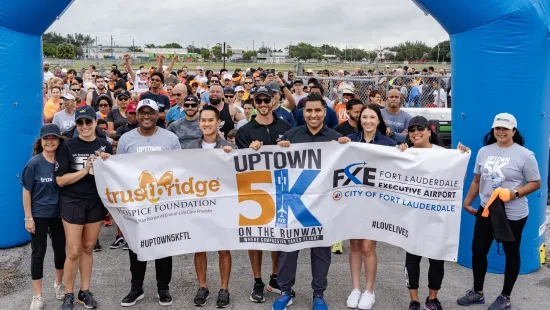 Diverse group holding Uptown 5K race banner at Fort Lauderdale Executive Airport starting line with large crowd behind.