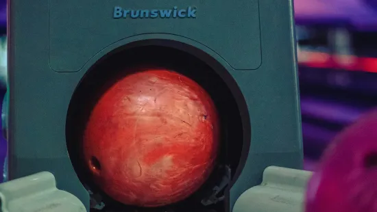 Red bowling ball resting in a Brunswick ball return rack at a bowling alley lane.