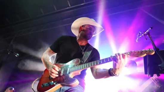 Musician in a white cowboy hat playing electric guitar on stage with vibrant purple and white lighting effects