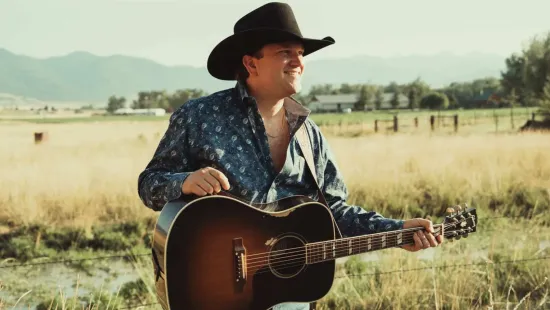 Man wearing cowboy hat playing acoustic guitar in an open field with mountains in the background