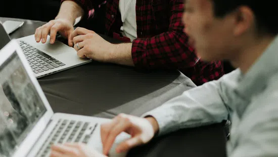 Two men collaborating and smiling while working on laptops at a table with a smartphone nearby