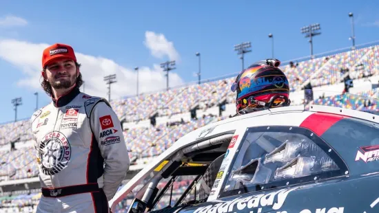 Race car driver standing next to a black and red stock car at a bright racetrack with empty grandstands.