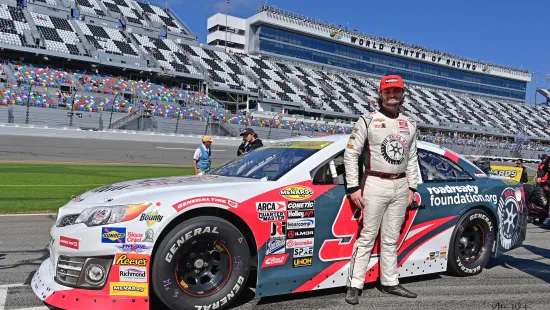 Race car driver in white suit stands next to red, white, and blue NASCAR car at Daytona racetrack with grandstands in background
